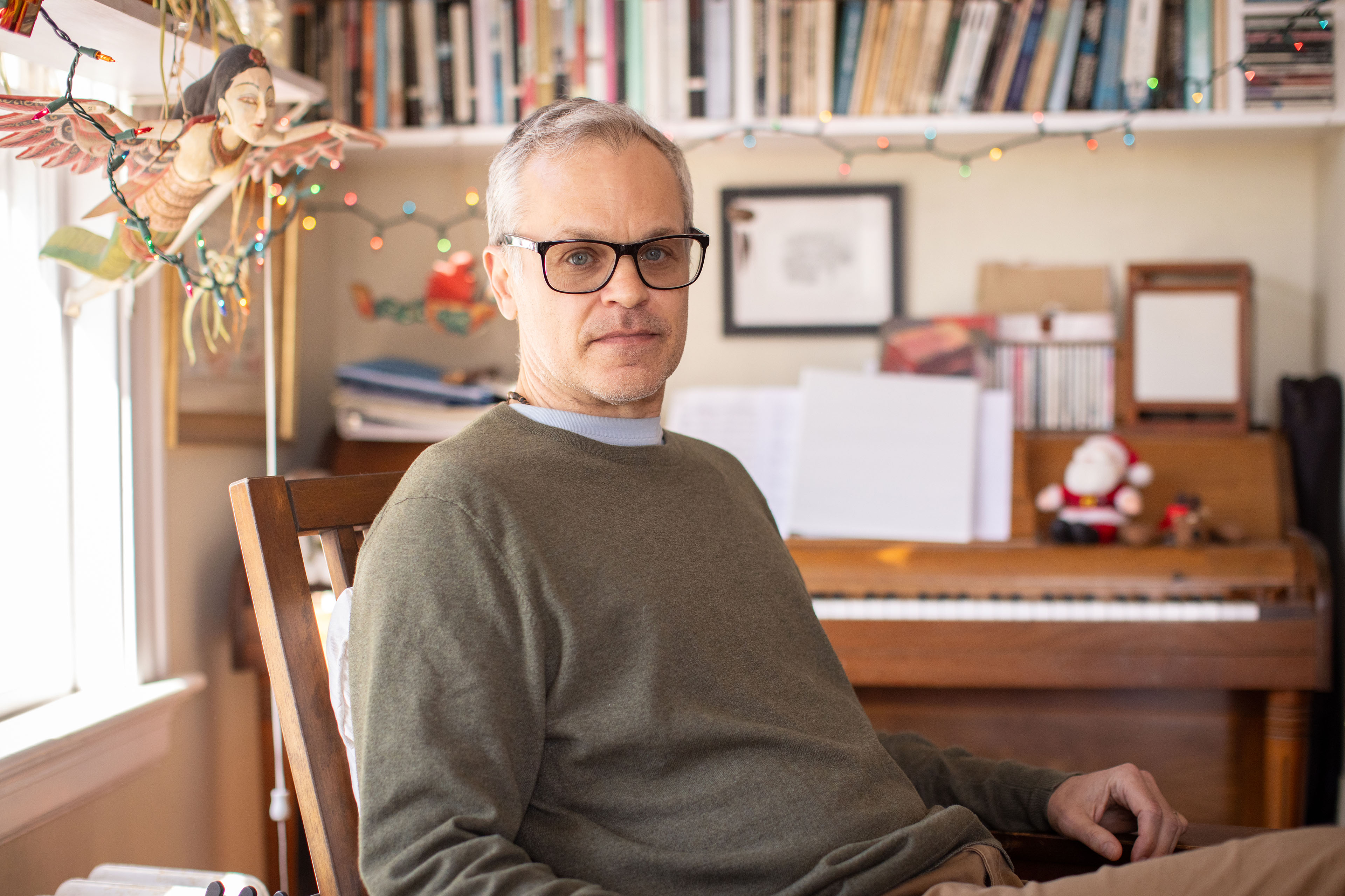 A man with gray hair and black-rimmed glasses poses for a portrait, sitting in a chair in front of a piano